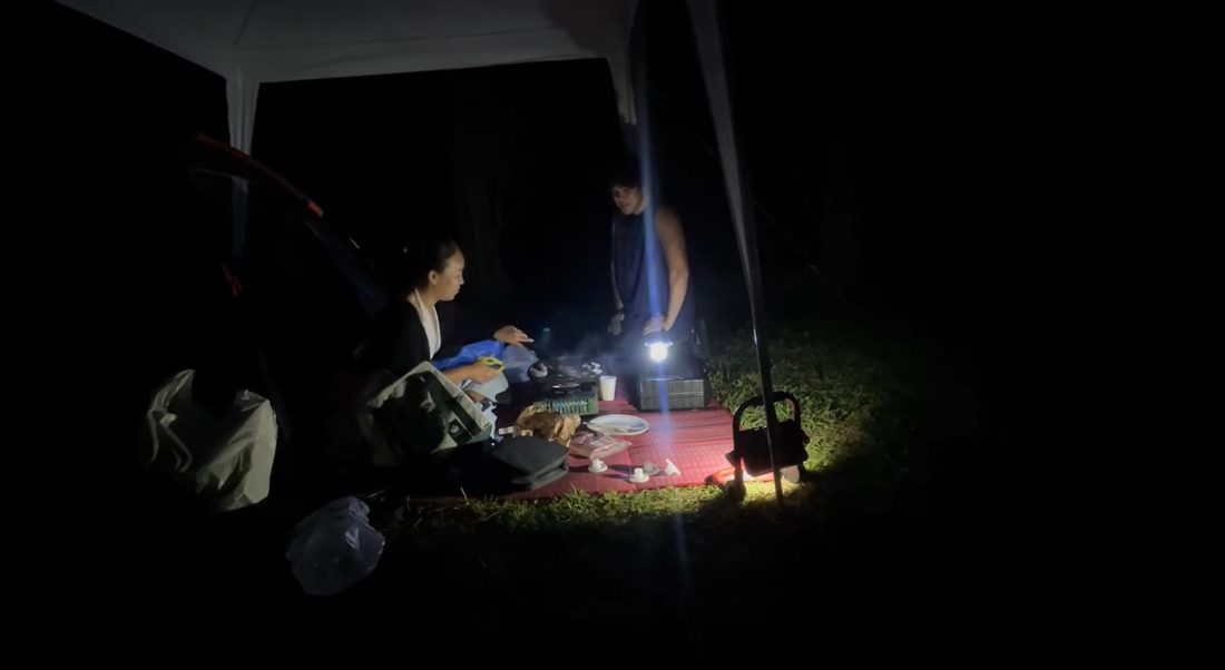 Night camping at Crosslands Reserve with two campers preparing food under a gazebo, using portable camping lights, a picnic mat and cooking gear after arriving in the dark.