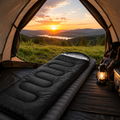 Black waterproof sleeping bag on an air mattress inside a camping tent at sunset with green grass and camping gear overlooking a scenic landscape.