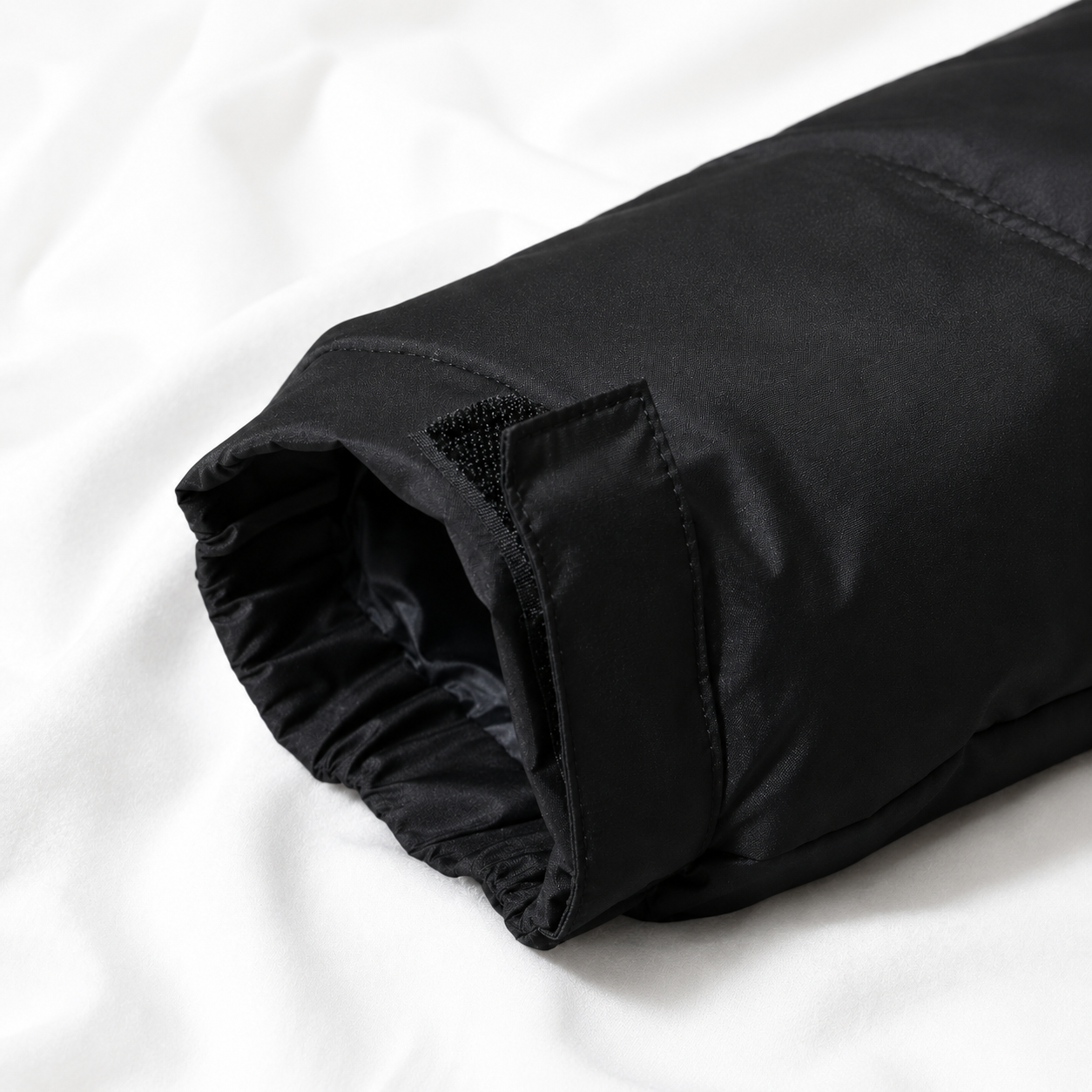 Close-up of a black puffer jacket sleeve with an adjustable Velcro cuff displayed on a soft white cloth background, highlighting the insulated design and weather-resistant finish.