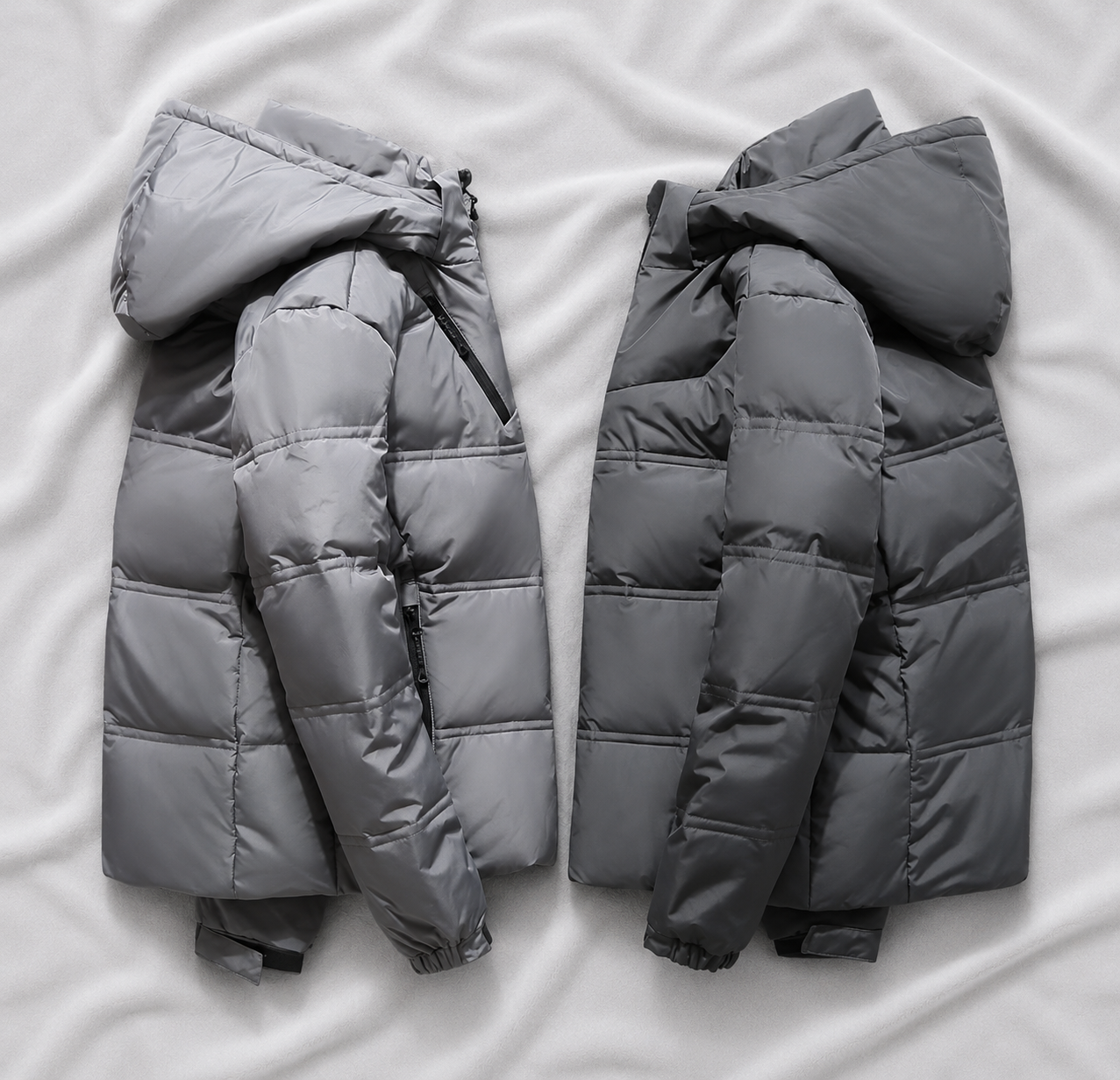 Light grey and dark grey hooded puffer jackets displayed on a soft light grey cloth background, showcasing insulated quilted construction and warm winter outerwear for cold-weather use.